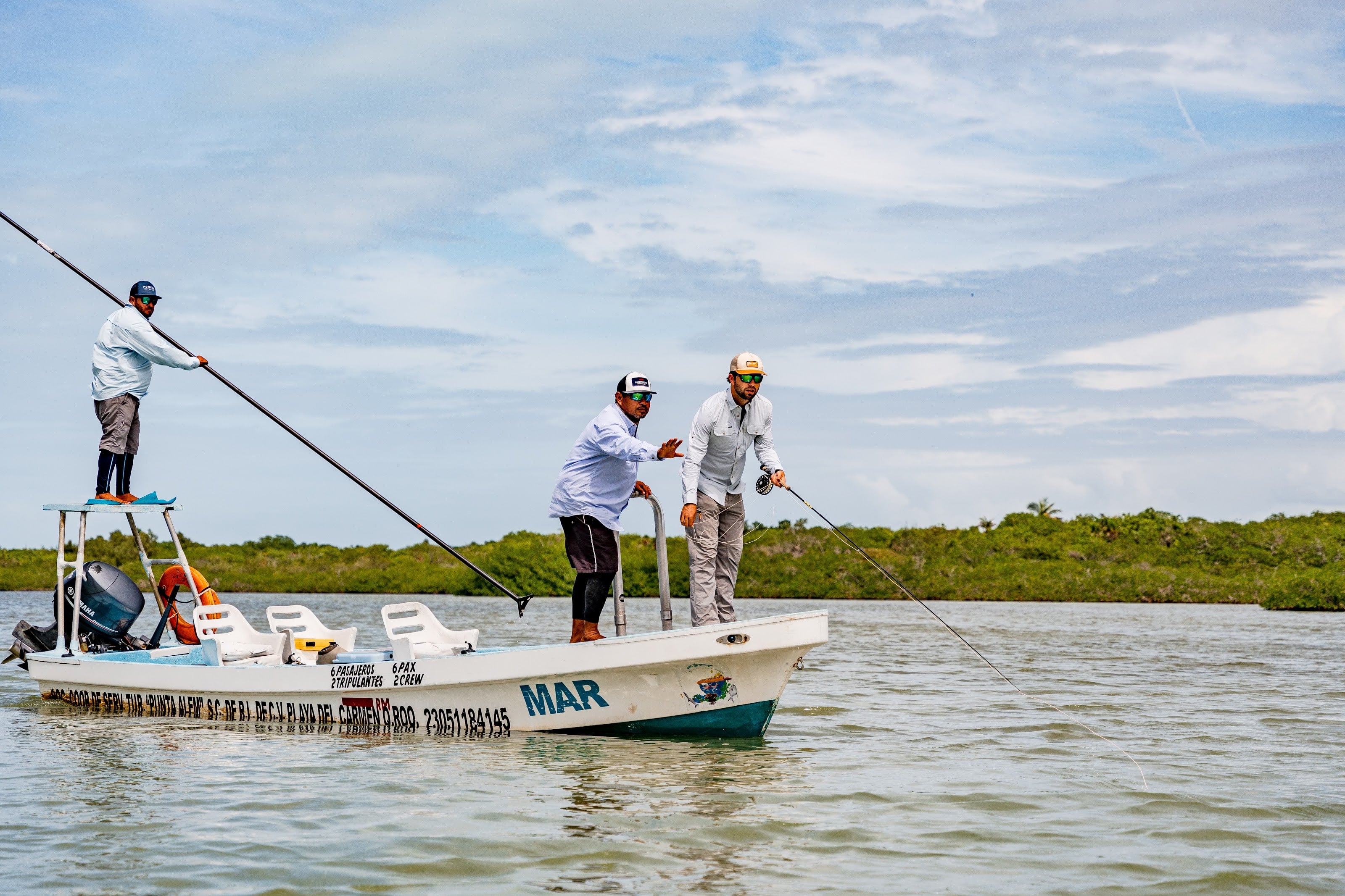 A guide poling a skiff across a mangrove flat while an angler waits on the bow